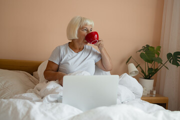 Caucasian senior 60s woman sitting with cup of coffee on bed and having breakfast while enjoying...