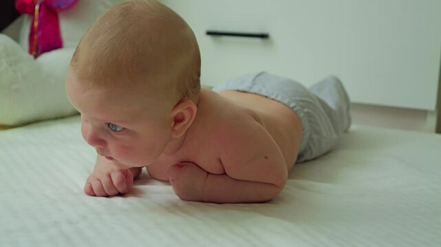 A baby with blue eyes and blond hair lying on his stomach. He is wearing grey trousers. The baby tries to crawl, but his head overbalances and falls on the sheet
