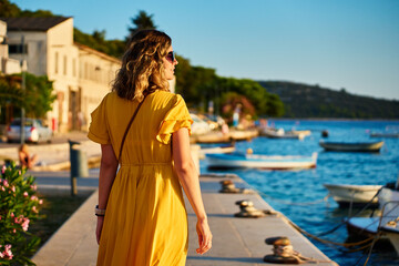 Fototapeta premium Woman is walking alone on sunny promenade with palm trees and flowers. Female tourist strolls along marina at sunset in coastal town