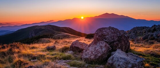  A sunset over mountain ranges, dotted with rocks in the foreground, and a lush, grassy field at their base