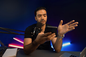 Broadcaster talking to listener on live streaming online, checking message from smartphone on social media in host channel by creative positive thinking concept at purple neon light studio. Surmise.