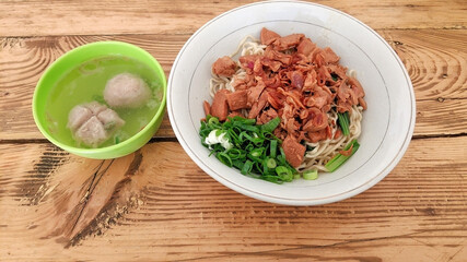 A traditional Indonesian chicken noodle dish served in a white bowl, topped with spring onions, chicken pieces and a bowl of meatball soup. The dish is served on a rustic wooden table.