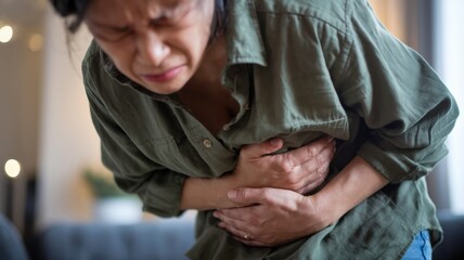 A photo of an adult holding their stomach and wincing in pain, with a blurred background. The adult is wearing a green shirt and is in a room. The lighting is soft.