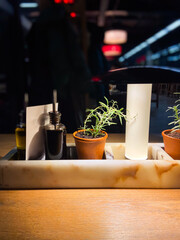 Cozy restaurant table setting with olive oil and balsamic vinegar bottles, flanked by small potted rosemary plants under warm lighting. Blurred background evokes a bustling yet intimate atmosphere