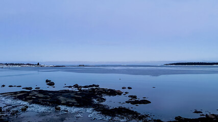 Calm winter seascape with icy shoreline, blue sky, and distant snowy treeline, evoking peaceful solitude and reflection