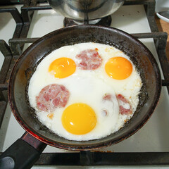 Fried eggs with pieces of sausage are fried on a gas stove in a black frying pan.