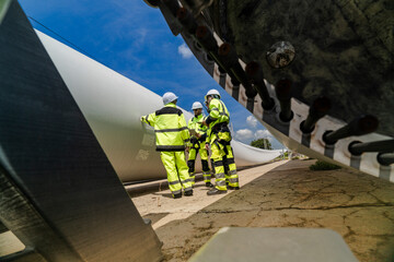 engineers in high visibility safety gear conduct a detailed inspection of a wind turbine blade at a construction site. The massive blade lays on the ground, highlighting renewable energy technology.