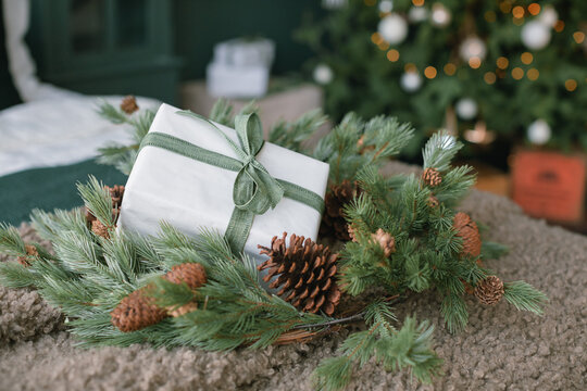 Close-up of a wrapped Christmas gift with a fir and pinecone wreath on a blanket in front of an illuminated Christmas tree