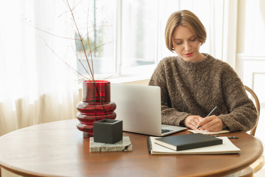 Young woman sitting at her dining table using her laptop and writing in a note pad