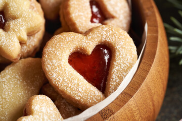 Closeup of a heart shaped traditonal Linzer Christmas cookie filled with red strawberry marmalade in a wooden bowl