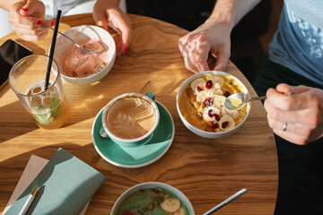 Overhead view of two people eating smoothie yogurt bowls with banana, cranberries, pumpkin seeds, coffee and juice