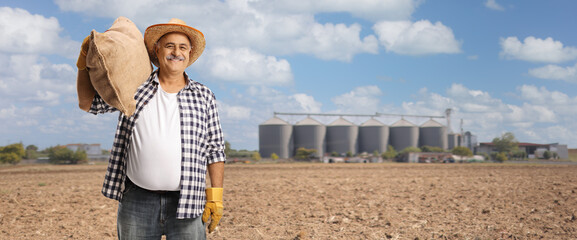 Mature farmer carrying a burlap sack on his shoulder on a field with tower silos © Ljupco Smokovski