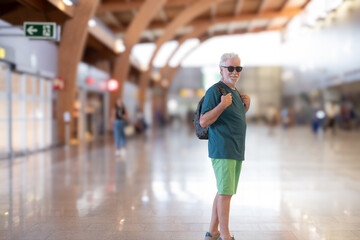 Handsome carefree senior man travels at airport with backpack walking towards board gate. Portrait of elderly happy traveler enjoying trip and vacation