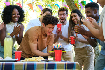 Friends are surprising a man holding a birthday cake with burning candles during a party in a park