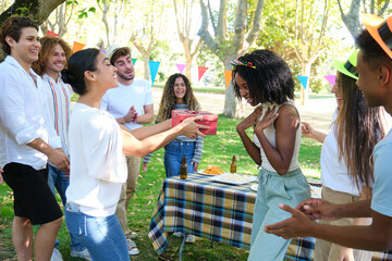 Group of friends are celebrating a birthday in a park, with one woman receiving a gift while her...