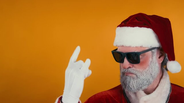 Stab closeup portrait of cheeky Santa Claus in sunglasses and traditional costume making horn hand sign and shaking head to upbeat rock music, smiling on yellow background. Template, copy space