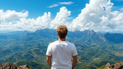 A young man stands on a rocky overlook, admiring the expansive mountain range. The scenery features lush greenery, distant peaks, and a brilliant blue sky, creating a serene atmosphere