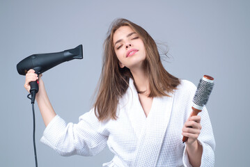 Woman in bathrobe combing hair, drying hair with hairdryer. Portrait of female model with a comb brushing hair. Girl with hairs brush and blow dryer. Hair care and beauty. Morning routine.