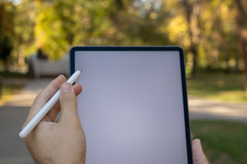 Young college boy using blank screen tablet with stylus pen while sitting outdoors in the park.