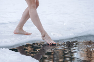 close-up view of girl legs standing on snow near ice hole getting into cold water in winter,...