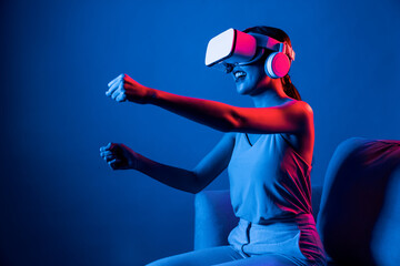 Smart Female sitting on sofa surrounded by neon light wear VR headset connecting metaverse,...