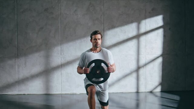 Strong sportsman doing lunges holding weight plate at light studio. Guy workout