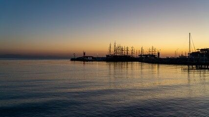 Fototapeta premium Sunrise at Kemer yacht marina in Antalya, Turkiye. The calm sea reflects the orange sky, capturing the serene beauty of this popular summer vacation spot.