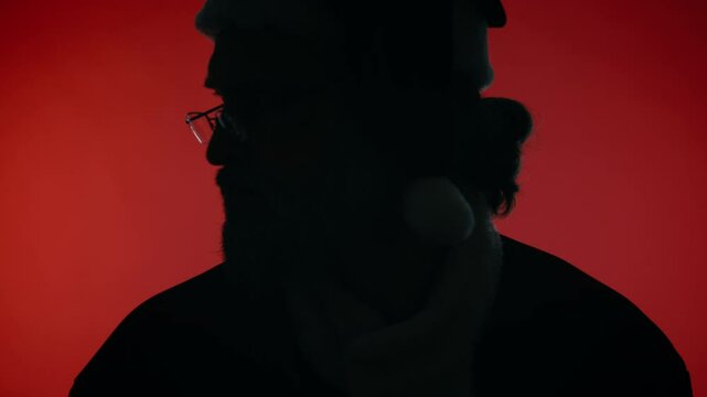 Stab closeup of dark silhouette of Santa Claus in glasses and costume passing by against red background and looking back, walking sneakily. Suitable for template, copy space