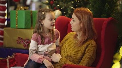 Medium shot of a young mother and her cute little daughter gossiping while sitting on sofa in the background of Christmas arrangement - Powered by Adobe