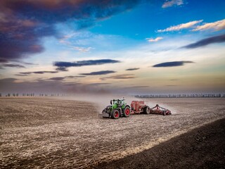 modern green tractor sowing wheat seeds in dusty field on sunset with colourful autumn sky 