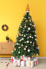 Decorated Christmas tree with gift boxes, silver balls, Santa hat and wooden cabinet near yellow wall