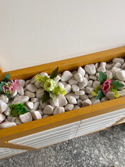 Wooden planter filled with white pebbles and colorful artificial flowers decorates an interior...