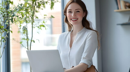 綺麗なオフィスの窓辺でノートパソコンを持つ美しい女性　パソコンを抱え笑顔　左にコピースペースあり