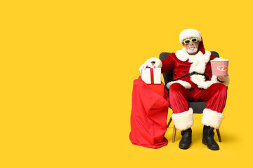 Santa Claus holding bucket of tasty popcorn and bag with gifts on yellow background