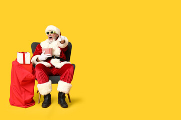 Shocked Santa Claus holding bucket of tasty popcorn and bag with gifts on yellow background