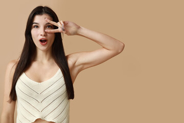 Surprised young woman showing peace sign on beige background