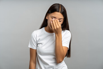 Weary, emotionally drained teenage girl rubs tired eyes, slouched against isolated gray wall. Girl...