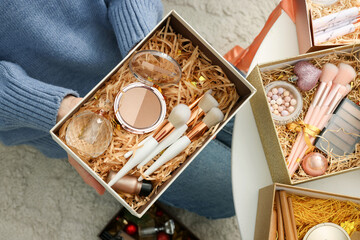Woman holding Christmas gift boxes with makeup brushes and cosmetics at white table, above view