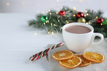 Yummy cocoa in cup, candy cane, dried orange slices and Christmas wreath on white wooden table. Space for text