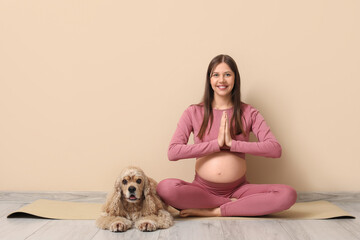 Young sporty pregnant woman with cocker spaniel meditating near beige wall
