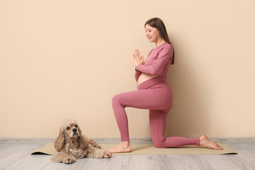 Young sporty pregnant woman with cocker spaniel meditating near beige wall