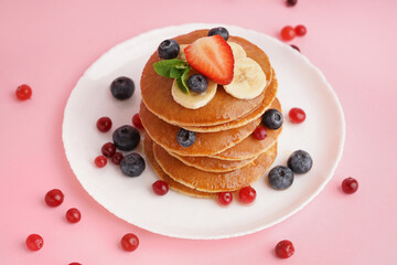 Plate with tasty pancakes, banana slices, cranberries and blueberries on pink background