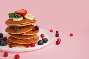 Plate with tasty pancakes, banana slices, cranberries and blueberries on pink background
