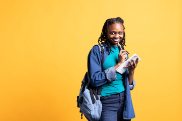 Portrait of upbeat BIPOC pupil smiling, reading and writing for school exam on notepad, isolated...
