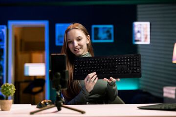 Cheerful teenager presenting wireless keyboard from brand sponsoring video, urging fans to purchase it, filming with phone. Happy gen Z girl does influencer marketing for computer peripheral product