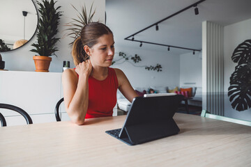 Woman in red dress sitting at a table with a tablet, holding her neck in discomfort. Looking to laptop screen too much, neck hurts.