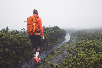 A smiling woman in an orange puffy jacket stands by a misty lake surrounded by greenery. She wears a headband and has a relaxed posture, enjoying the serene outdoor environment. The fog creates a tran