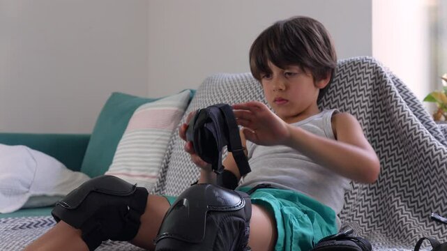 Young boy putting on elbow pads while seated on the couch, securing the protective gear in place. He is focused on tightening the strap, preparing for a sports activity
