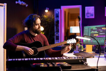 Songwriter trying to tune the strings of his guitar in home studio, adjusting knobs before playing...