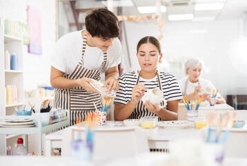 Positive enthusiastic guy assisting interested young girl painting ceramic mugs and plates in pottery class. Creative hobby concept..